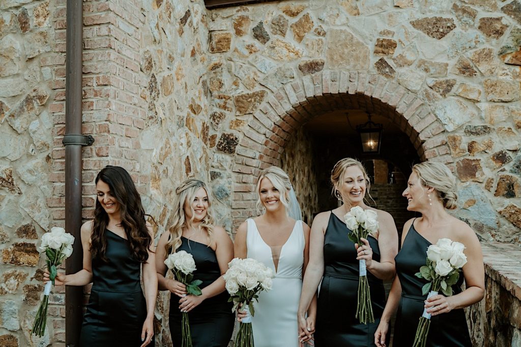 A bride stands with her 4 bridesmaids, 2 on either side of her, all dressed for the wedding and holding bouquets of white roses underneath a stone archway at their intimate Florida wedding venue
