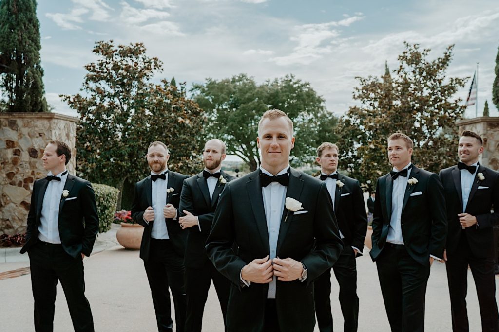 A groom stands with his 6 groomsmen, 3 on either side of him all dressed for their wedding, outside of their intimate Florida wedding venue. The groom smiles and looks at the camera while the others look in various directions.