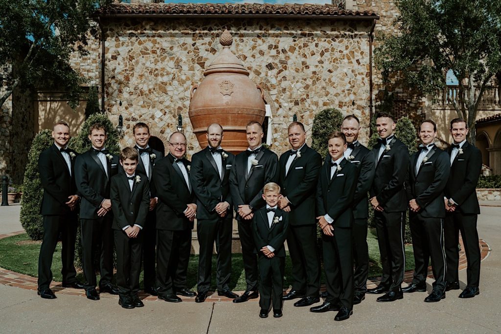 A groom stands with his groomsmen and male family members outside of their intimate Florida wedding venue all dressed in black tuxedos for the wedding.