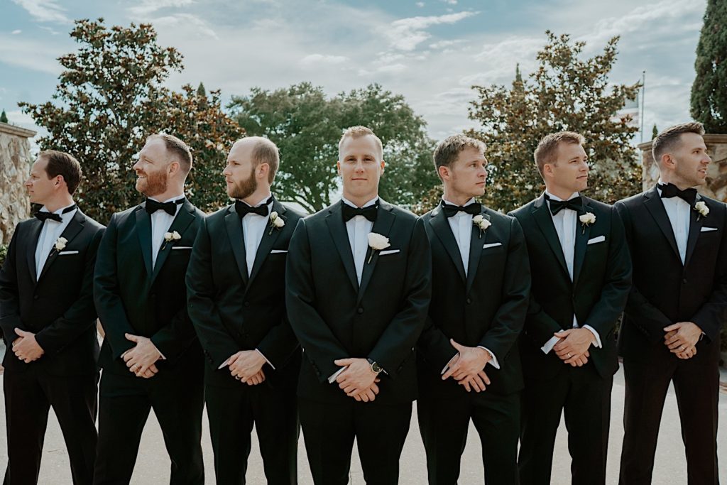 A groom stands with his 6 groomsmen, 3 on either side of him all dressed for their wedding, outside of their intimate Florida wedding venue. The groom smiles and looks at the camera while the men on the left look left and the men on the right look right
