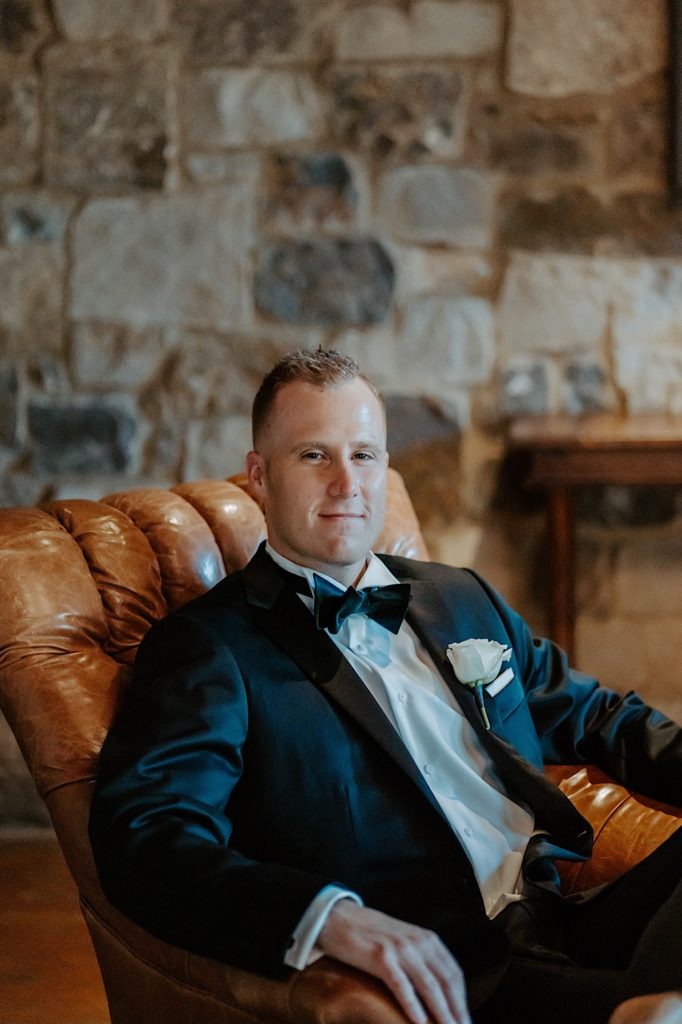 A groom dressed for his wedding sits in a brown leather chair and smiles at the camera