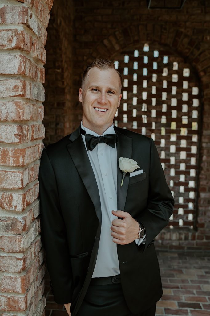 A groom dressed for his wedding stands and smiles at the camera while leaning against a brick wall.