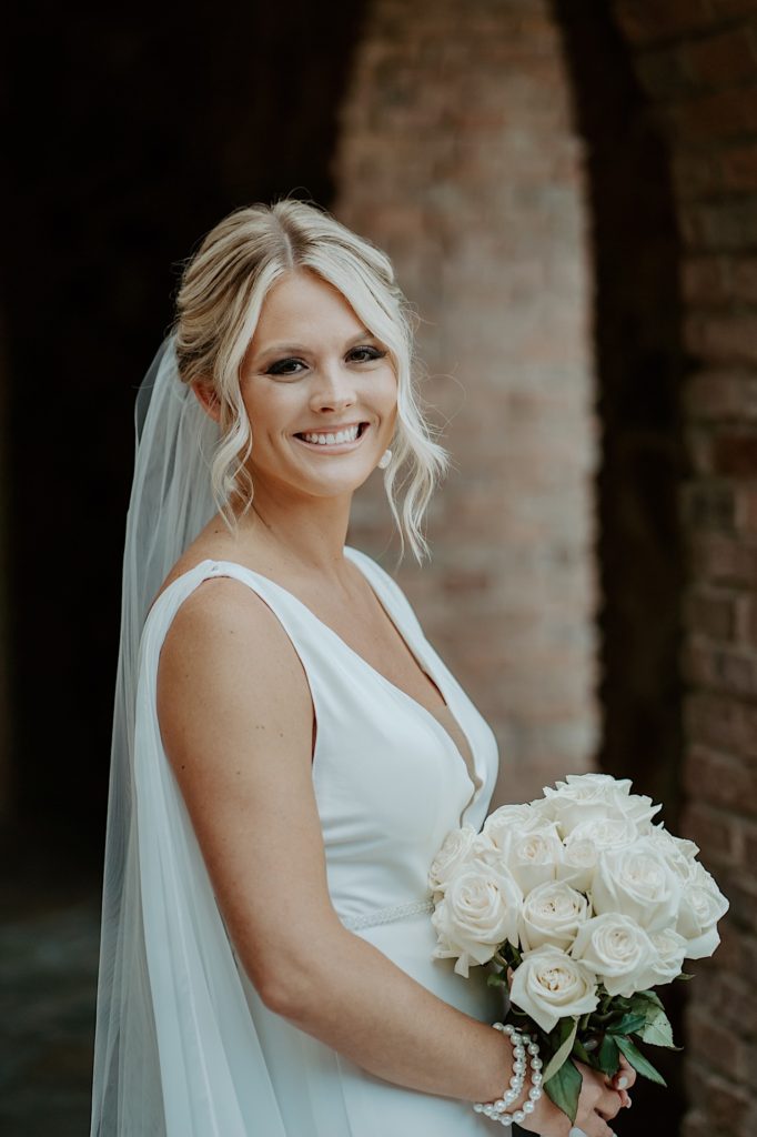 Portrait of a bride smiling at the camera wearing her wedding dress and holding a bouquet of white roses