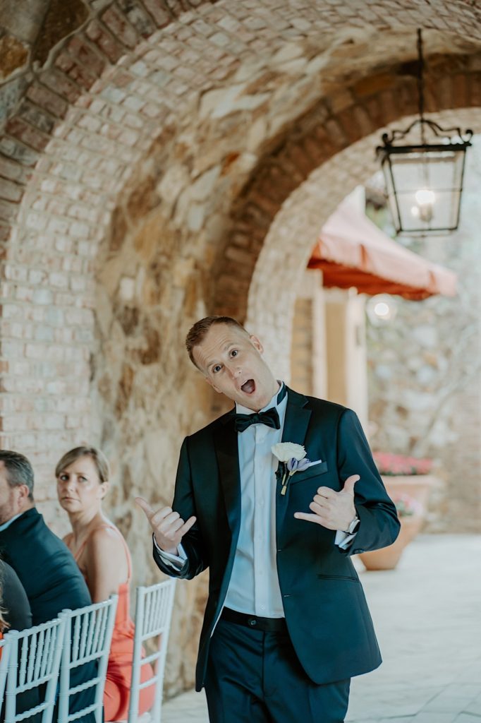 The groom of a wedding walks towards the camera under a stone archway while gesturing at the camera with guests to his left.