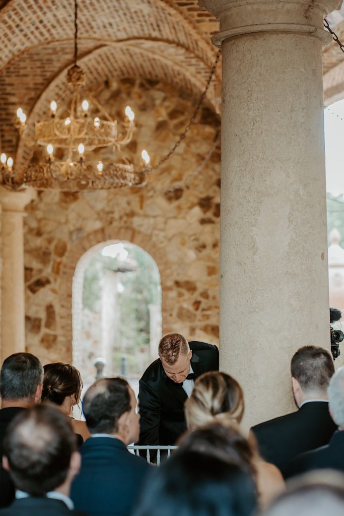 Photo of a groom leaning over to adjust a chair during his wedding ceremony with guests in the foreground