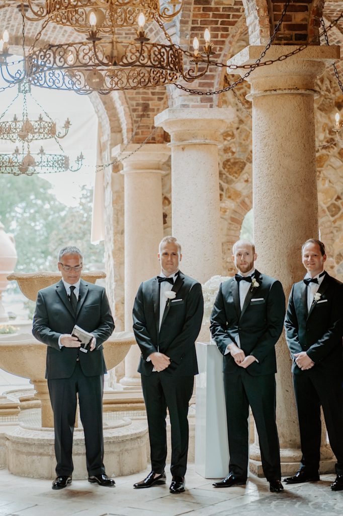 A groom stands with two groomsmen to his right and the officiant of the wedding to his left as they wait for the bride to enter the ceremony.