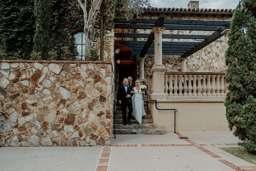 A bride walks down a stone staircase with her father to head towards their intimate Florida wedding ceremony