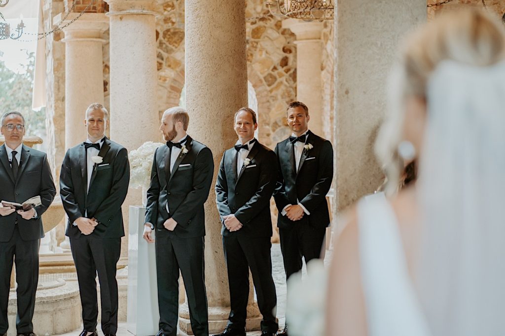 A groom smiles with his groomsmen next to him as he sees his bride in her wedding dress as she walks towards him for their wedding ceremony. The bride is out of focus in the foreground of the photo.