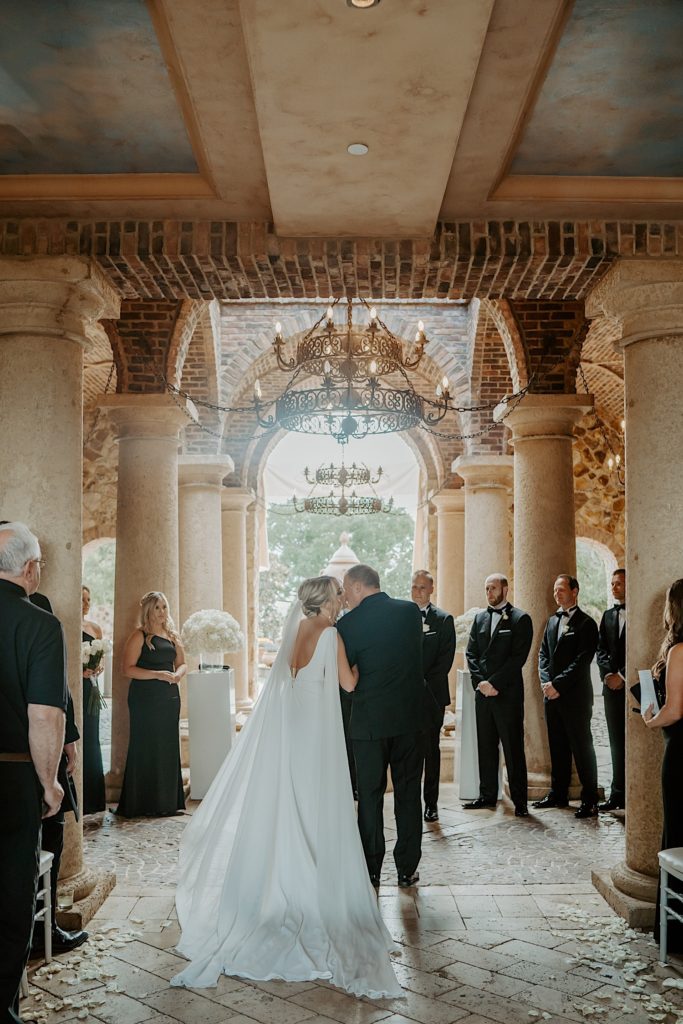 A bride walks down the aisle with her father facing away from the camera during her intimate Florida wedding ceremony and kisses him on the cheek.