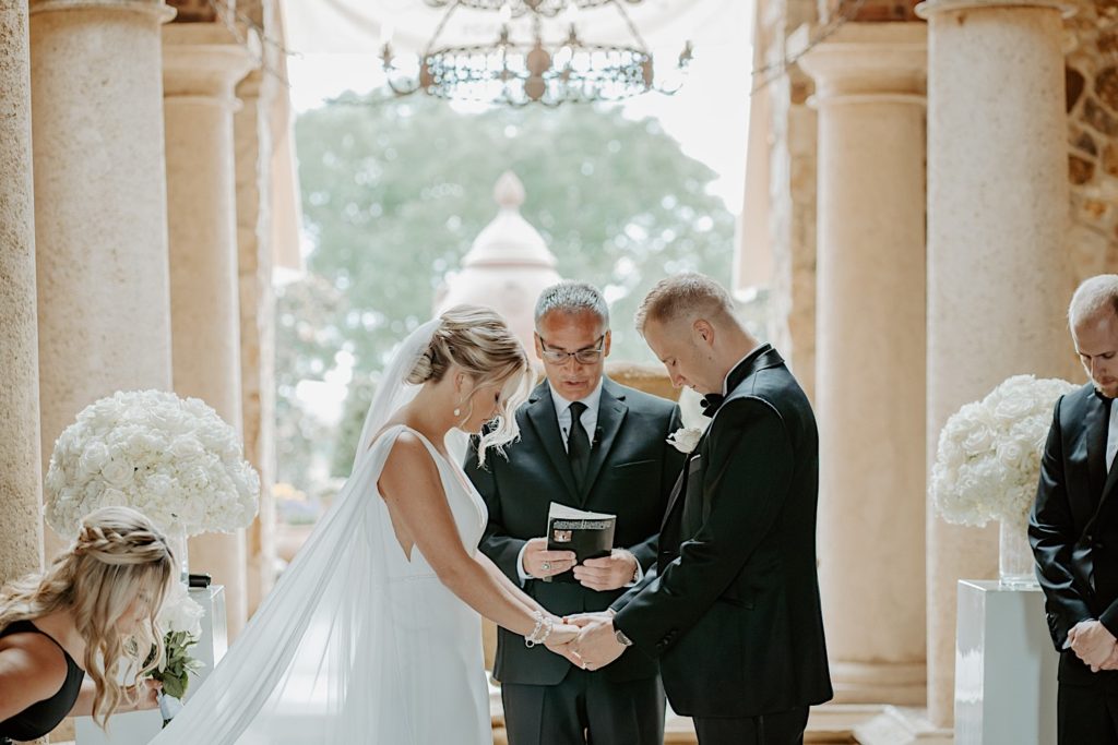 At an intimate wedding ceremony at Bella Collina in Florida a bride and groom both look down as they face each other holding hands with their officiant speaking behind them.