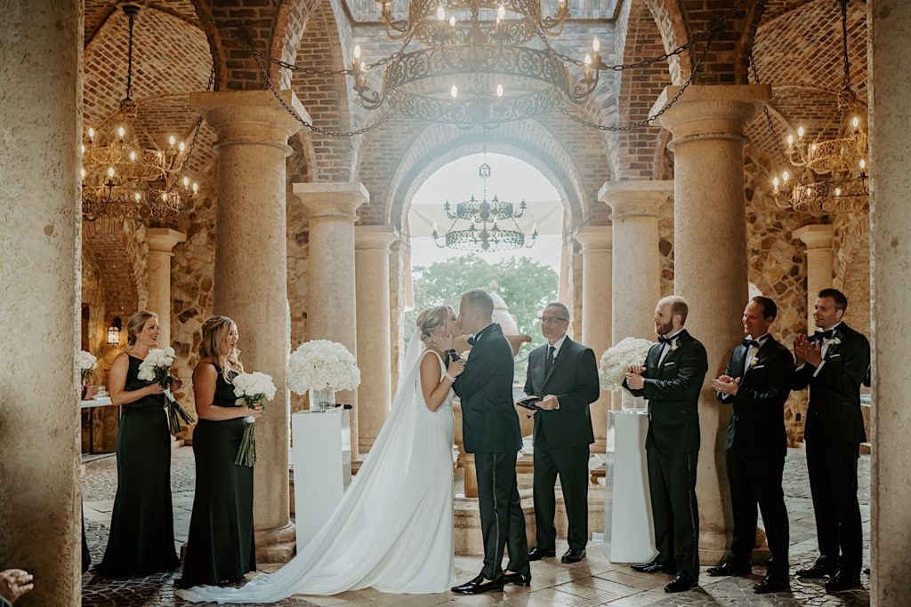 At an intimate wedding ceremony at Bella Collina in Florida a bride and groom kiss as their wedding parties clap and cheer them on
