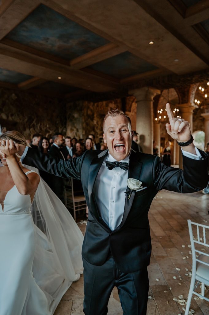 Photo of a groom cheering and smiling at the camera pointing his finger to the sky as he holds his brides hand while they exit their wedding ceremony.