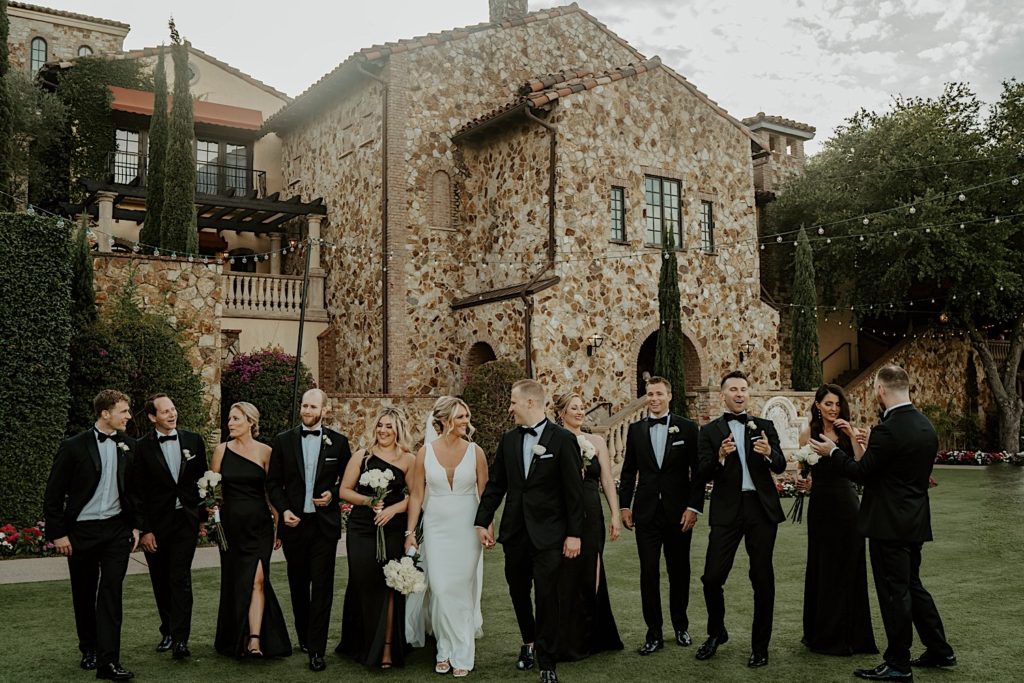 A bride and groom hold hands and walk towards the camera with their wedding parties to either side of them mingling, they're outside their intimate wedding venue Bella Collina in Florida.