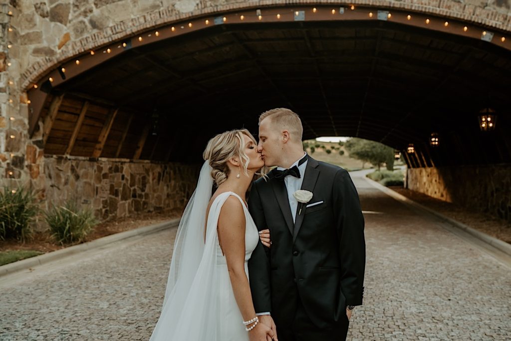 A bride and groom stand under a stone bridge and kiss one another after their intimate wedding ceremony at Bella Collina in Florida
