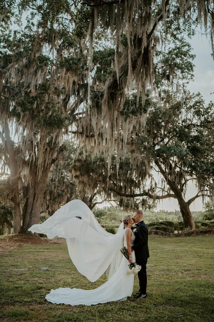 A bride and groom stand under large tree and kiss one another after their intimate wedding ceremony at Bella Collina in Florida while the bride's dress flows in the wind.
