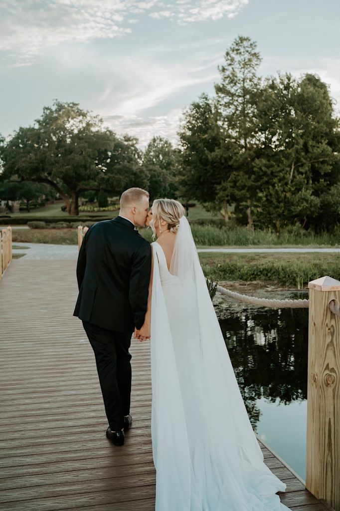 A bride and groom walk away from the camera and kiss while on a wooden bridge.