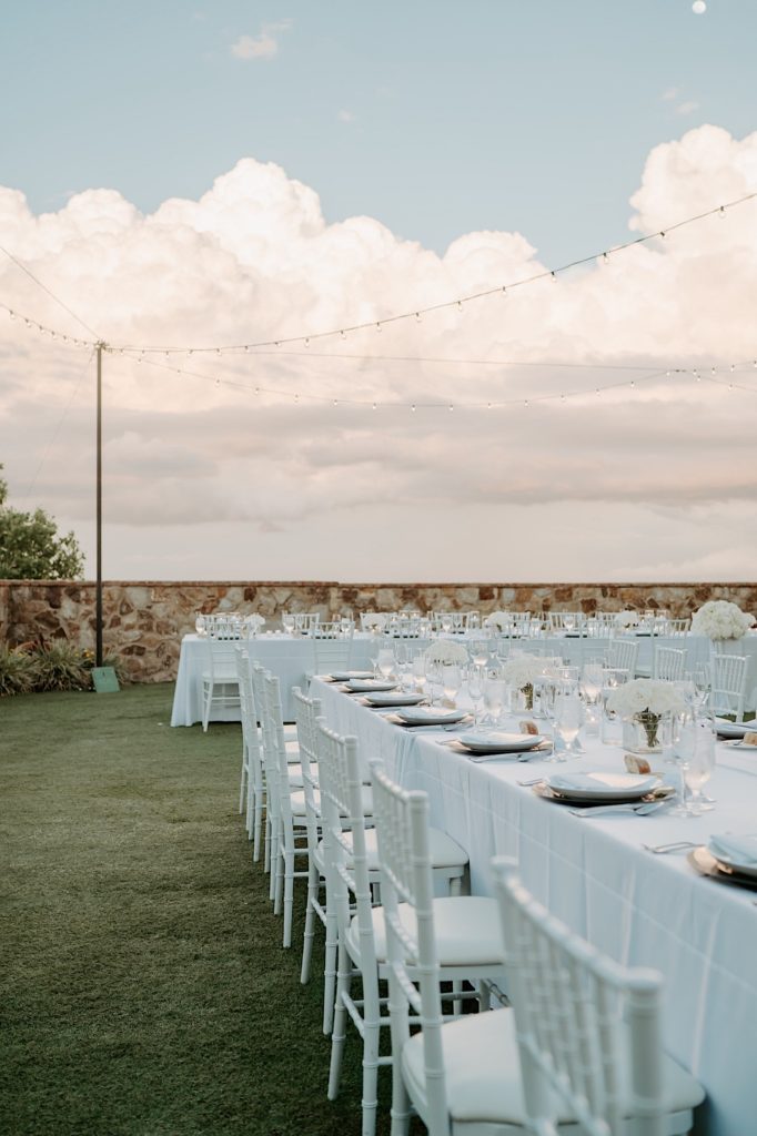 Tables set for an outdoor wedding reception with string lights above the tables and clouds in the background.