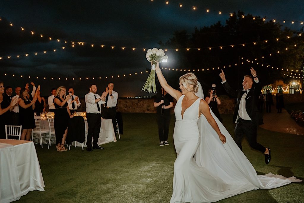 A bride and groom enter their intimate wedding reception at Bella Collina in Florida, the bride walks with her white rose bouquet in the air while the groom cheers behind her and their guests clap