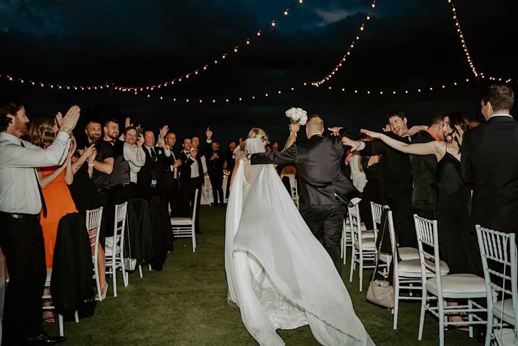 A bride and groom enter their intimate wedding reception at Bella Collina in Florida, the bride walks with her white rose bouquet in the air while their guests cheer around them.
