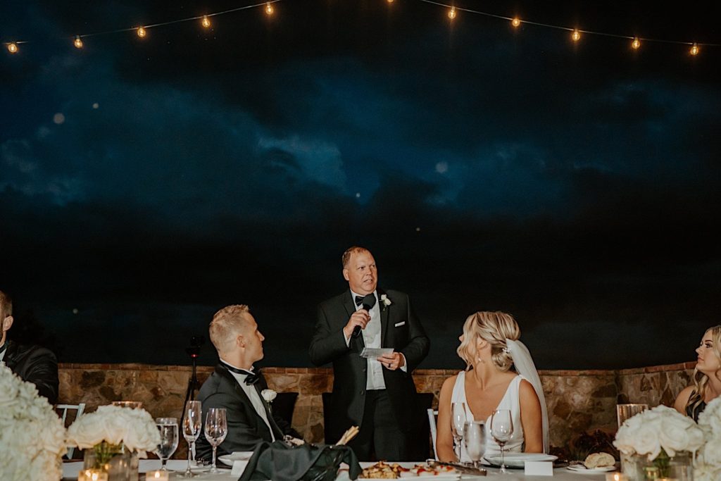A father stands between the bride and groom during their wedding reception and gives a speech, there are string lights and storm clouds overhead.