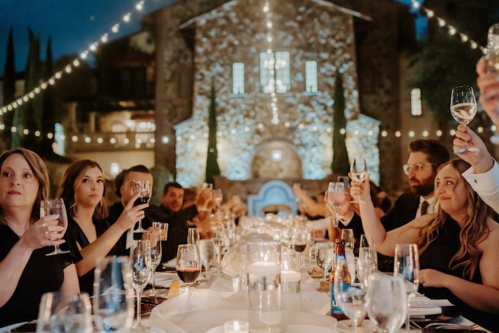Guests seated at a table set for an intimate wedding reception at Bella Collina in Florida raise their glasses during a speech.