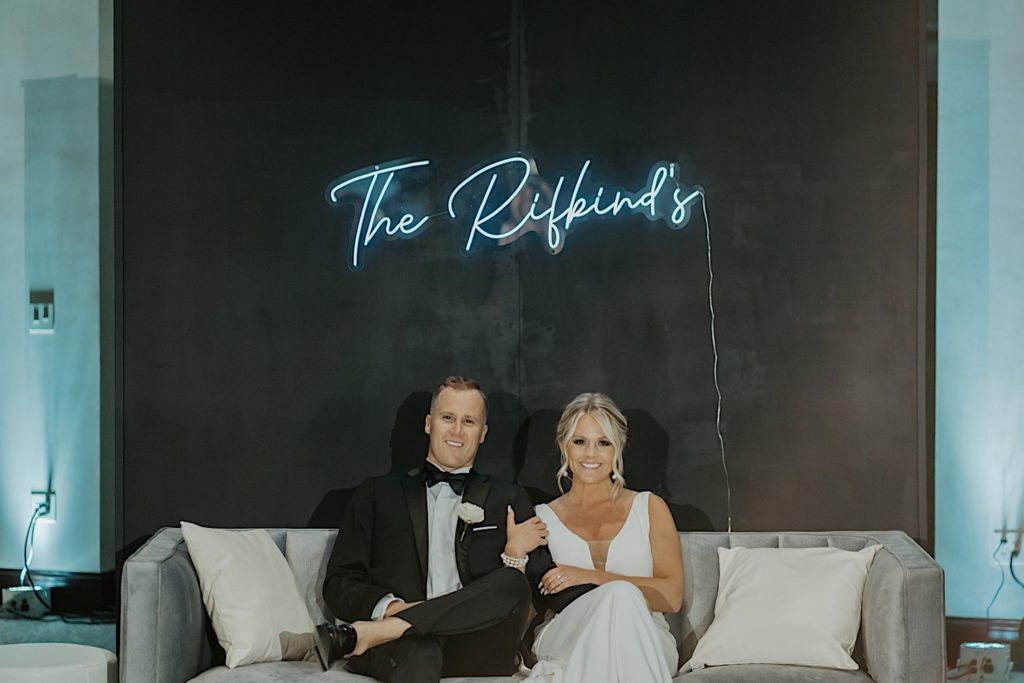A bride and groom sit on a couch together and smile at the camera with a neon sign of their last name on the wall behind them.