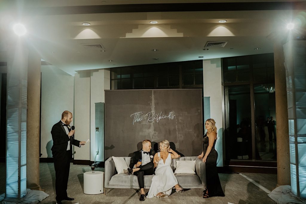 A bride and groom sit on a couch together and smile at one another while a groomsmen gives a speech to the left of them and a bridesmaid stands to the right of them.