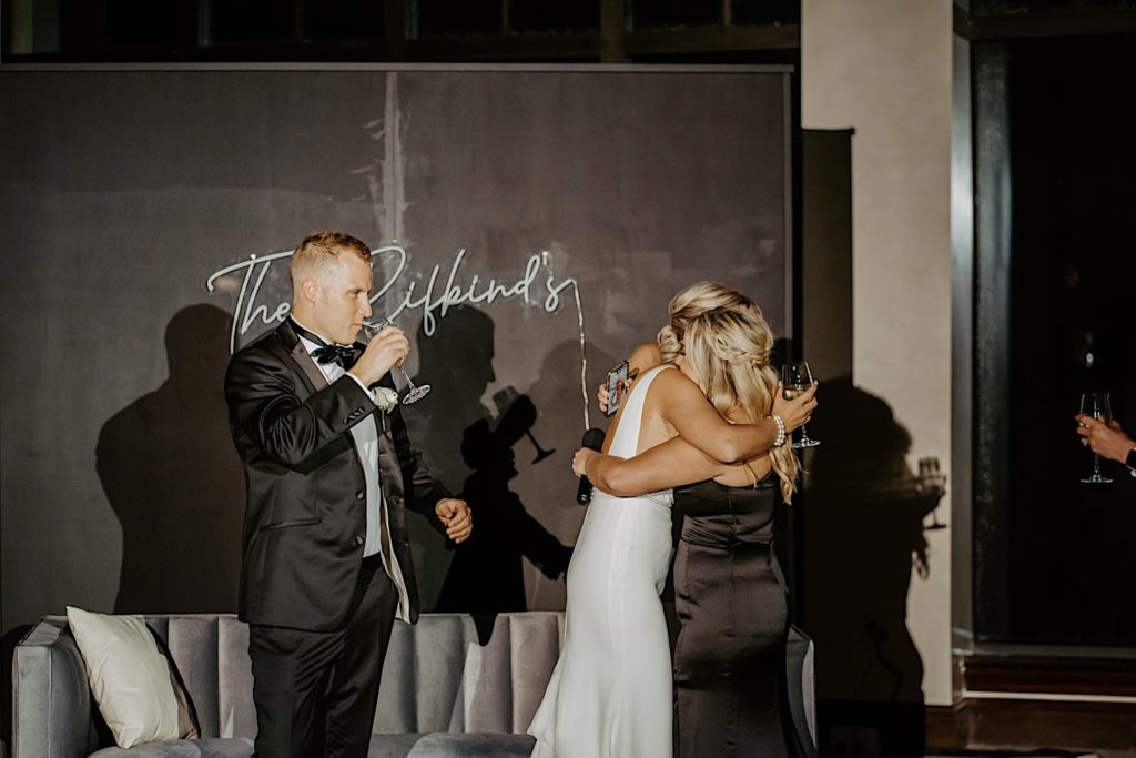A bride hugs one of her bridesmaids after a speech while the groom takes a sip of his drink to the left of them.