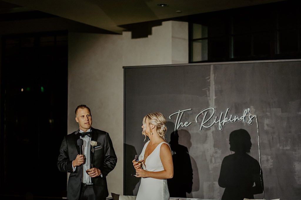 A bride and groom stand next to one another with a neon sign of their last name on the wall behind them. The groom has a microphone and is giving a speech while the bride looks at him.