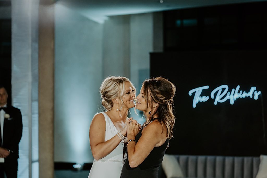 A bride and her bridesmaid smile at one another while dancing during a wedding reception with a neon sign of the bride's last name on the wall in the background.