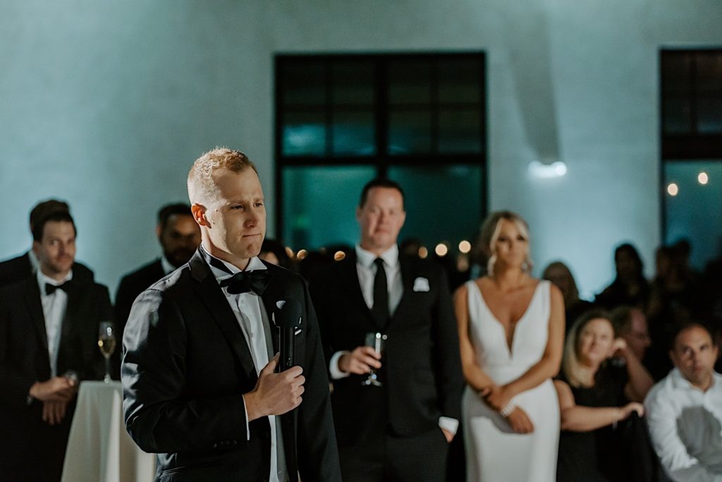 A groom stands holding a microphone during his wedding reception about to give a speech while his bride and the guests of their wedding are behind him.