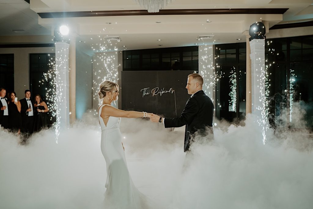 A bride and groom face one another and hold hands during their first dance at their intimate indoor wedding reception at Bella Collina in Florida. Small fireworks are lit on either side of them and and a smoke machine covers the floor in smoke.