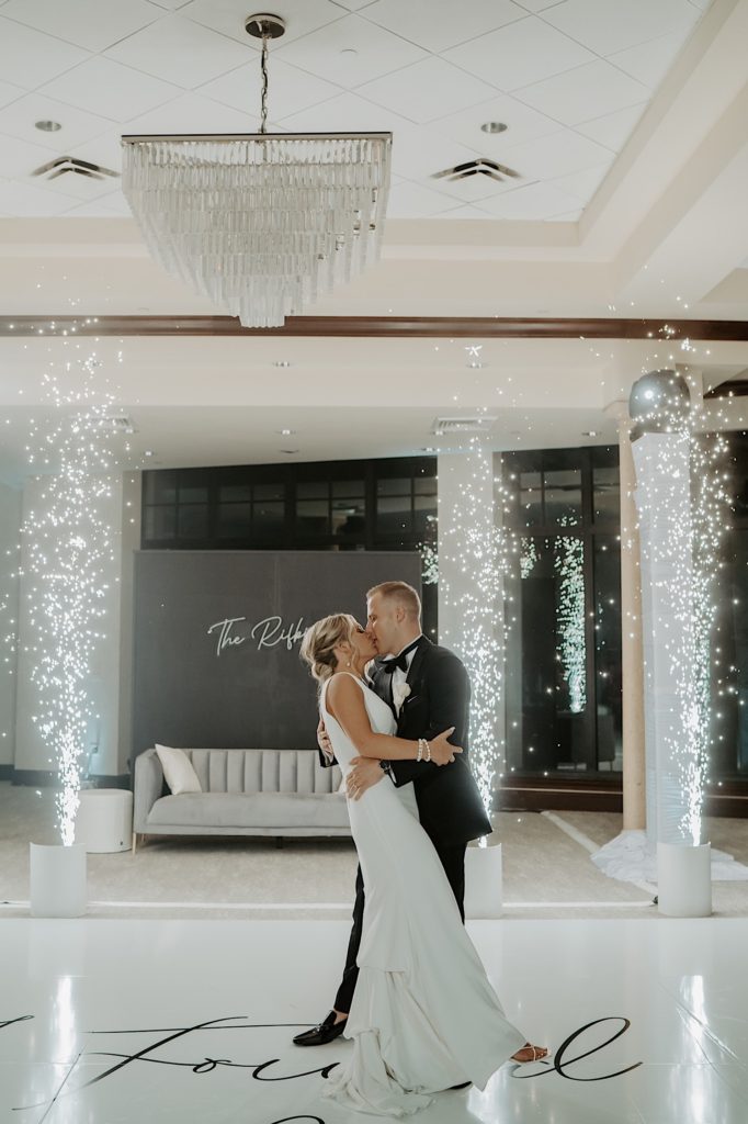 Photo of a bride and groom kissing during their intimate indoor wedding reception at Bella Collina in Florida. There are small fireworks lit on either side of them.
