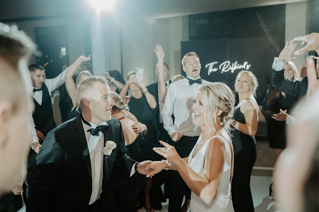 A bride and groom smile and dance with each other with guests surrounding them during their intimate wedding reception.