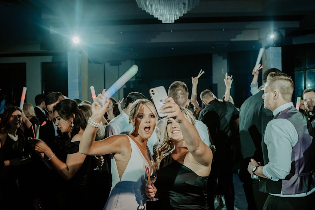 A bride poses with a bridesmaid for a selfie with guests dancing in the background during an intimate indoor wedding reception at Bella Collina in Florida.