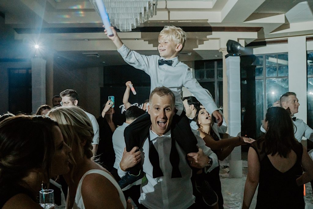 A young boy sits on the shoulders of the groom and waves a light up foam stick in the air during a wedding reception.