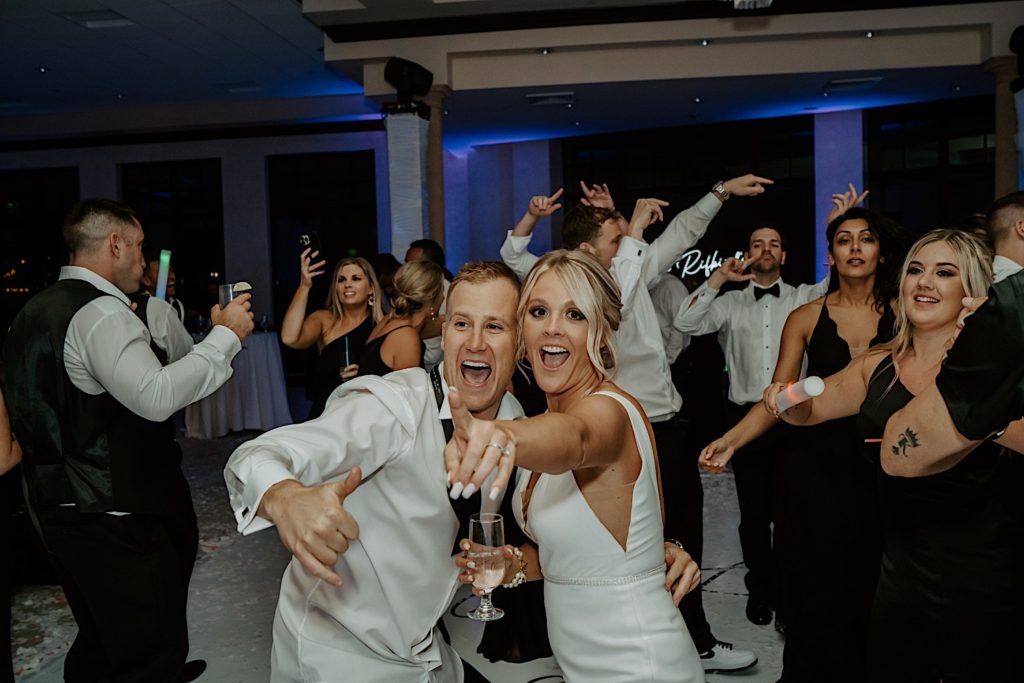 A bride points at the camera and smiles with the groom smiling next to her as they celebrate their wedding with guests dancing around them.