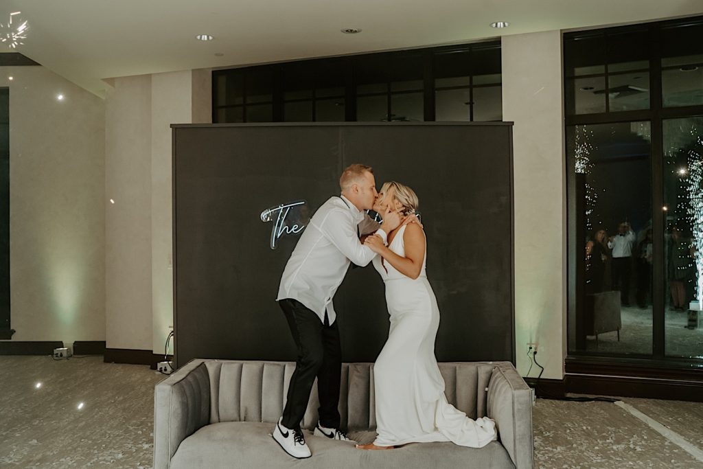 A bride and groom stand on a couch and kiss one another during their indoor wedding reception.
