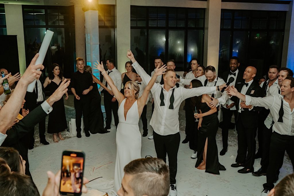 A bride and groom stand in the middle of their guests during their indoor wedding reception at Bella Collina in Florida. Their arms are extended into the air while their guests cheer around them.