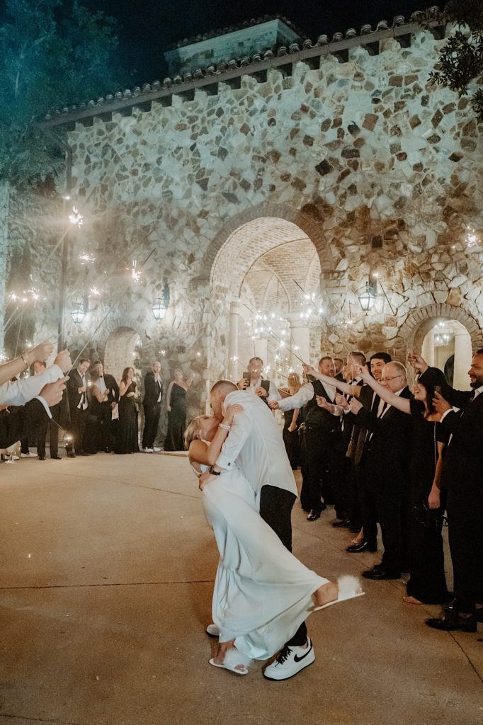 A groom dips his bride and kisses her with their guests around them holding sparklers as they exit their intimate wedding reception at Bella Collina in Florida.