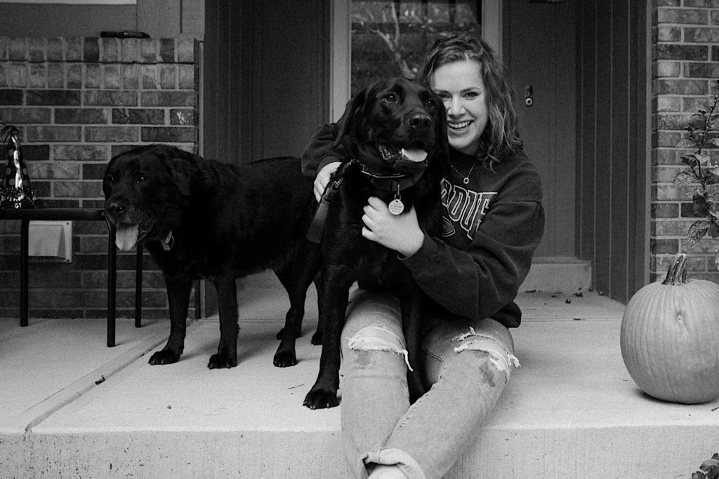 Black and white photograph of Shelby Jane Photography, an Indiana elopement photographer, smiling at the camera with her two black labs standing next to her