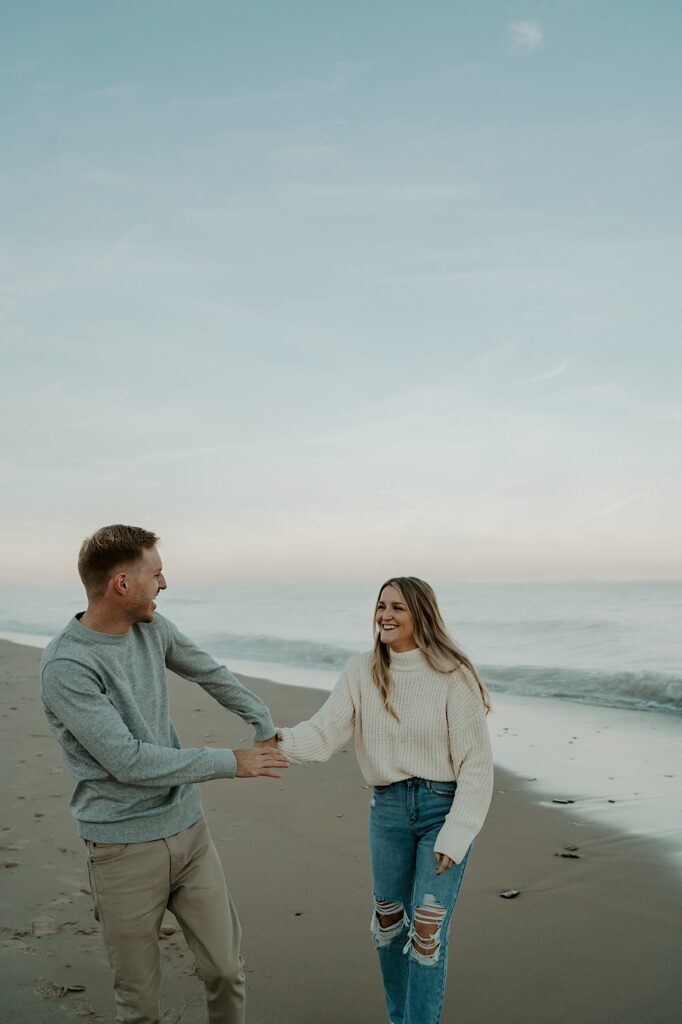 A couple hold hands and laughs with one another while walking towards the camera during their engagement session at the Indiana Dunes