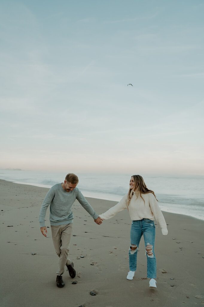 A couple holds hands during their engagement session walking towards the camera.  The fiancé wears khakis with a grey sweater and the soon to be bride wears ripped jeans with white tennis shoes and a cream turtle neck.