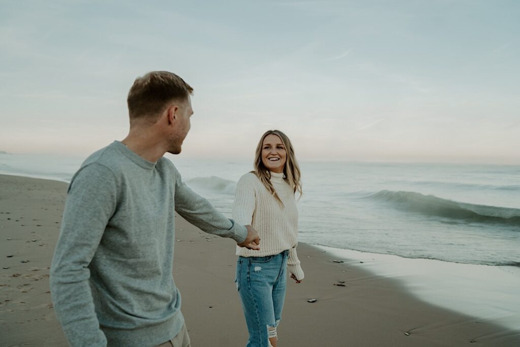 A couple holds hands on the beach during their fall engagement session at the Indiana Dunes.