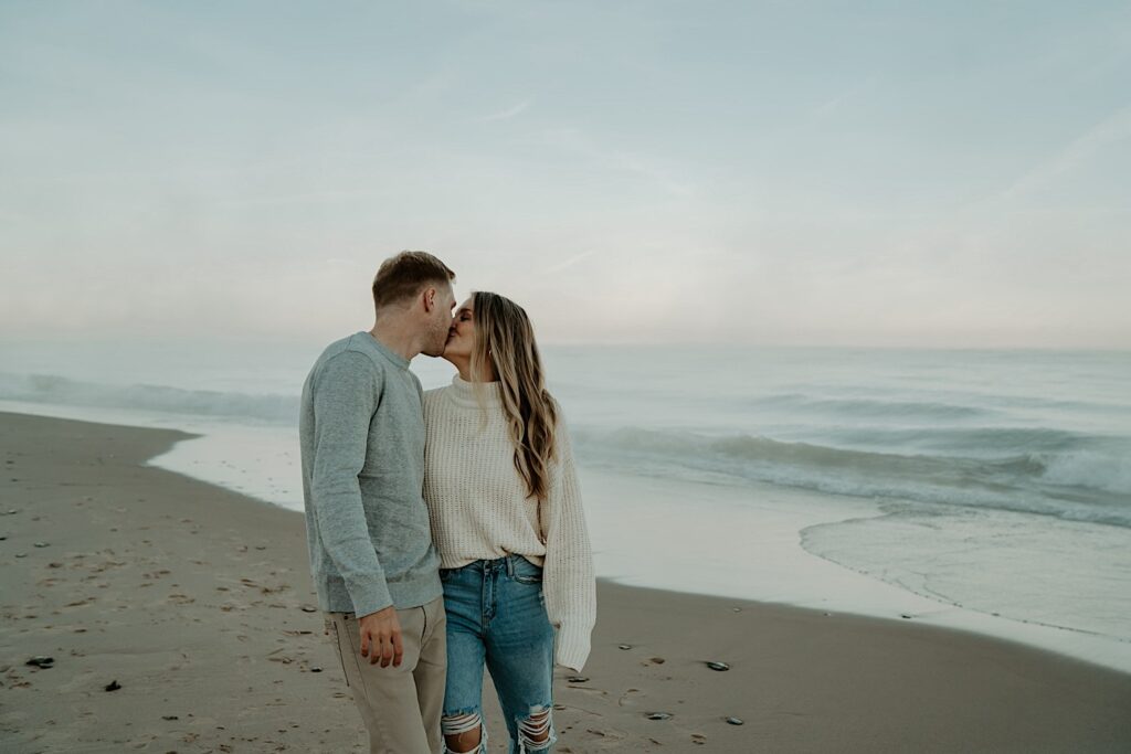 An engaged couple kisses while wearing cozy fall outfits during their engagement session at the Indiana Dunes during sunset.