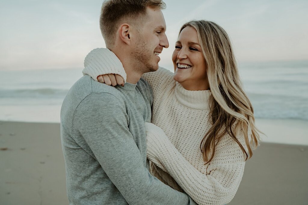 A fiancé hugs his fiancée while they laugh and look at one another.  They are both wearing cozy long sleeve shirts for their fall engagement session at the Indiana Dunes.