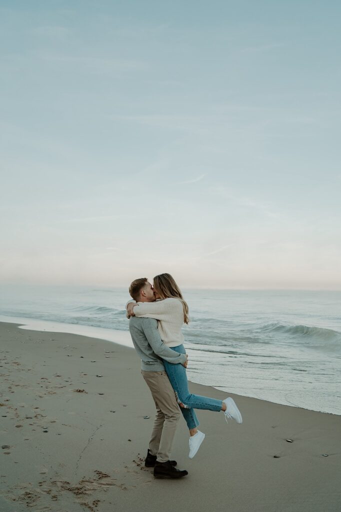 A fiancé holds his fiancée up while she wraps her arms around his shoulders and kisses him as they spin on the beach.