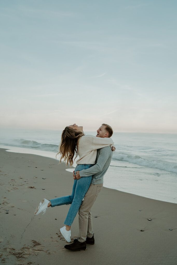 A fiancé holds his fiancée up while she wraps her arms around his shoulders and laughs as they dance and spin on the beach.