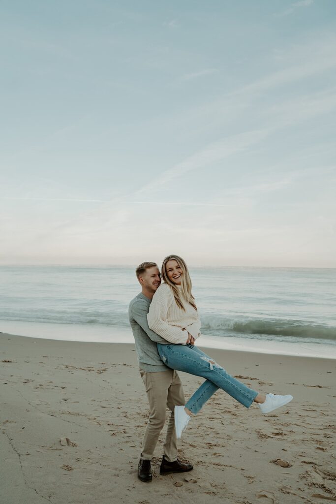 A fiancé holds his soon to be wife and spins her around, while she laughs.  They are on the beach watching the sunset during their engagement session.