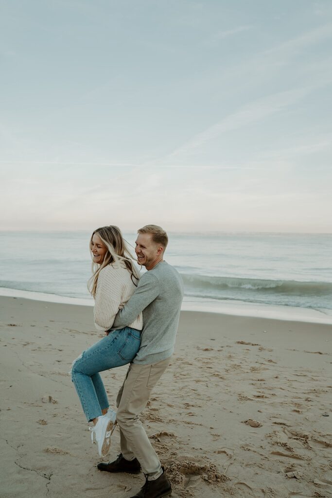 A fiancé holds his soon to be wife and spins her around, while she laughs.  They are on the beach watching the sunset during their engagement session.